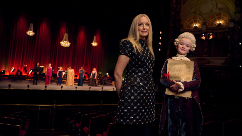 A woman in a black dress stands beside a child dressed in an 18th-century costume holding sheet music in a theatre, with a stage and performers blurred in the background.