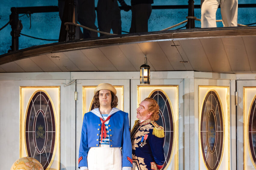 Two actors in nautical-themed costumes stand on stage; one in a blue sailor outfit and another in a navy uniform. A sailor stands on an upper deck, while three figures are silhouetted against a blue backdrop.