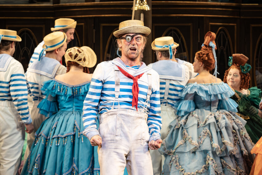 A man in a striped sailor shirt and straw hat, wearing a monocle and red neckerchief, performs expressively on stage. Behind him, people in ornate blue and teal period costumes stand in a group.