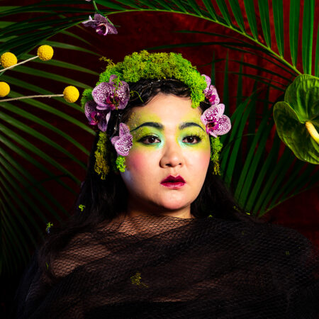 A person draped in black mesh poses against a vivid backdrop of green palm leaves and red. They wear green make-up and a headdress of moss, purple flowers, and greenery, creating a dramatic, nature-inspired look.