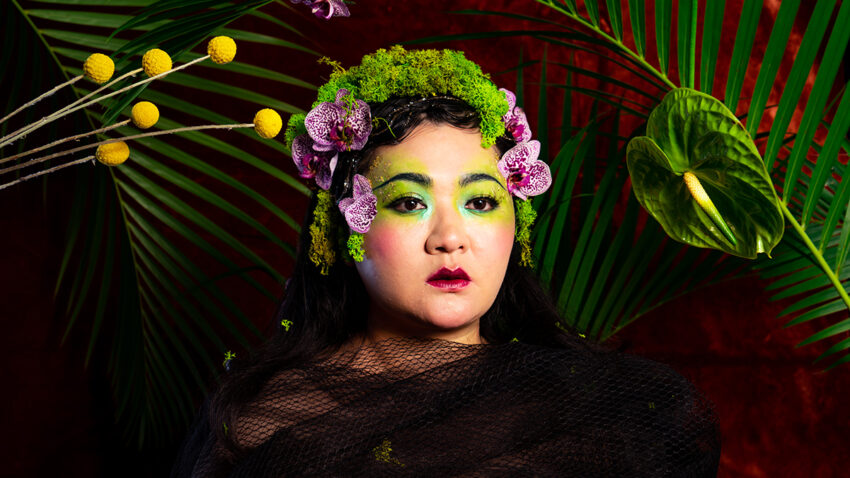 A person draped in black mesh poses against a vivid backdrop of green palm leaves and red. They wear green make-up and a headdress of moss, purple flowers, and greenery, creating a dramatic, nature-inspired look.