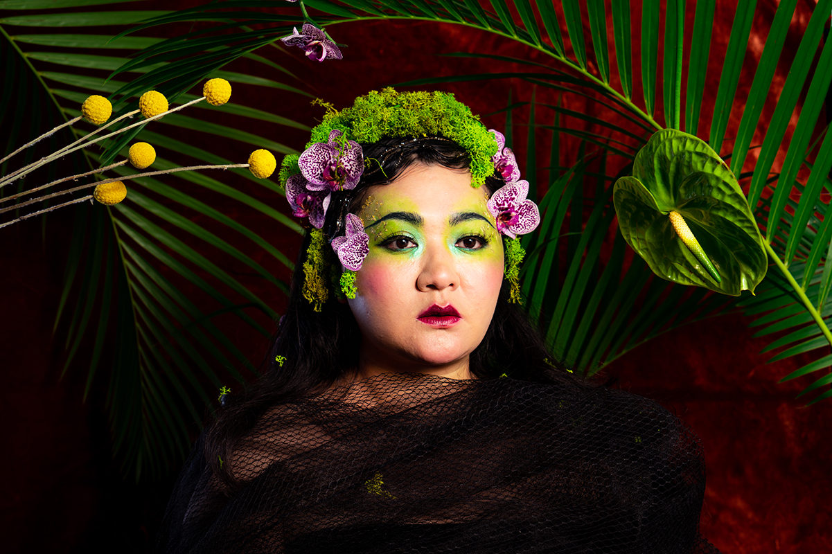 A person draped in black mesh poses against a vivid backdrop of green palm leaves and red. They wear green make-up and a headdress of moss, purple flowers, and greenery, creating a dramatic, nature-inspired look.