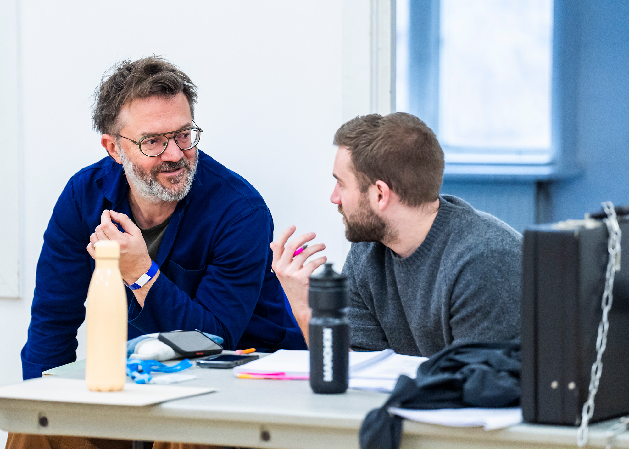 Two men sit at a table having a lively conversation. Papers, water bottles, and a briefcase are on the table. The man on the left wears glasses and a blue shirt; the man on the right wears a grey jumper.