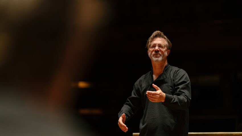 André de Ridder conducts an orchestra, standing behind a music stand and gesturing with his hands. He wears a black shirt and appears focused, with a blurred figure in the foreground.