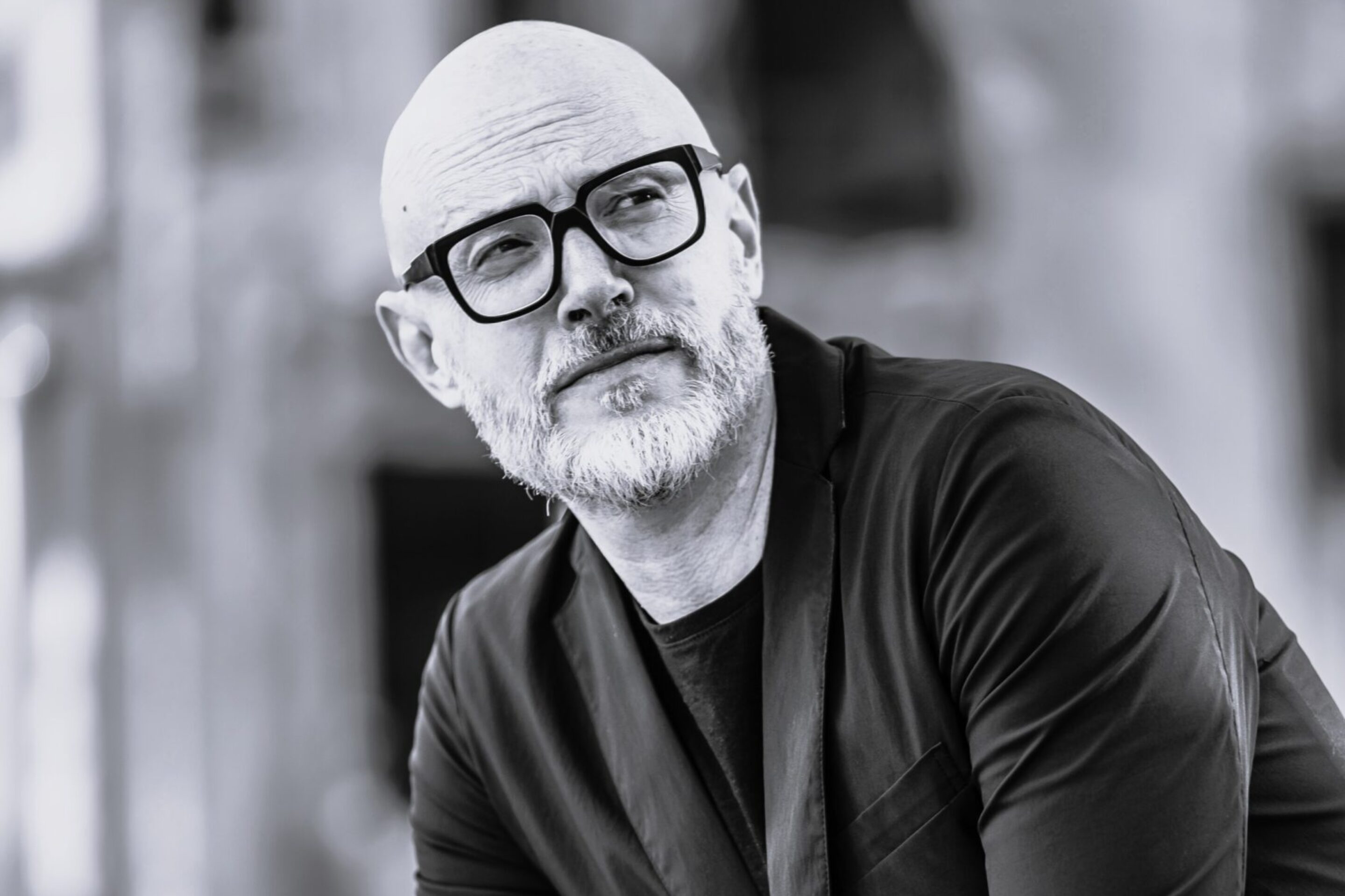 Baldur Brönnimann, a middle-aged man with a beard and glasses, dressed in a black blazer and jeans, poses thoughtfully outdoors. The photo is in black and white, with a blurred background.