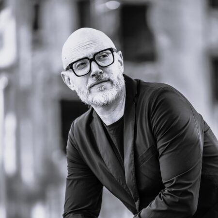 Baldur Brönnimann, a middle-aged man with a beard and glasses, dressed in a black blazer and jeans, poses thoughtfully outdoors. The photo is in black and white, with a blurred background.