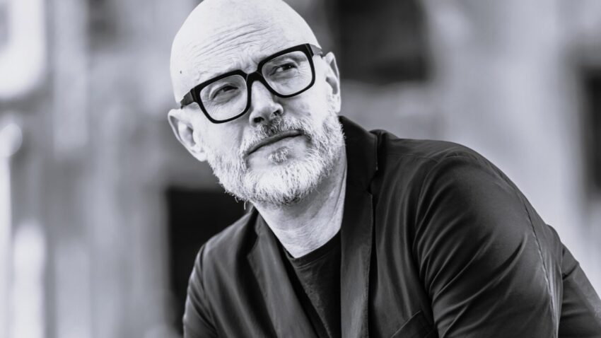Baldur Brönnimann, a middle-aged man with a beard and glasses, dressed in a black blazer and jeans, poses thoughtfully outdoors. The photo is in black and white, with a blurred background.