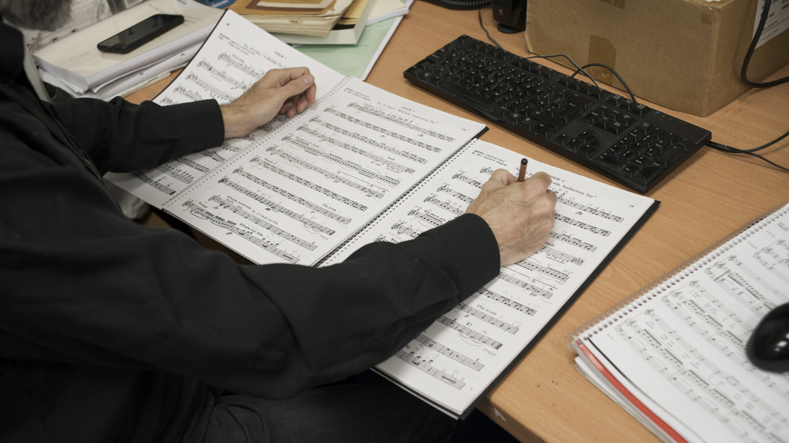 A person writes on manuscript paper at a desk, surrounded by a computer keyboard, papers, and folders—capturing the creative spirit that drives initiatives like the ENO Annual Appeal. The focus is on their hands, musical notes, and workspace.