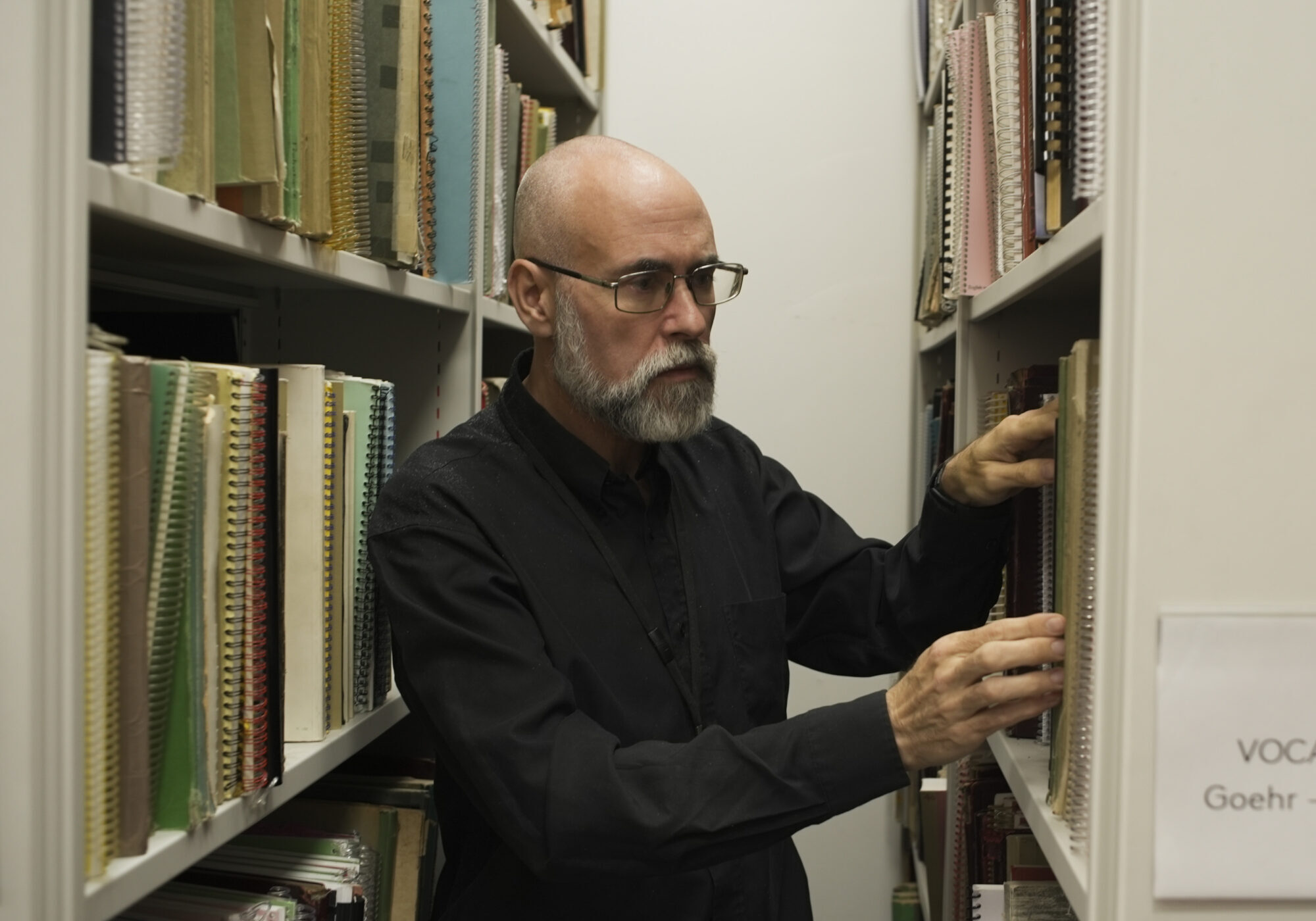A man with glasses and a grey beard stands between bookshelves, reaching for a book. A sign on the shelf reads Vocal Scores Goehr - Mussorgsky, part of the ENO Annual Appeal collection. Shelves are filled with various books and scores.