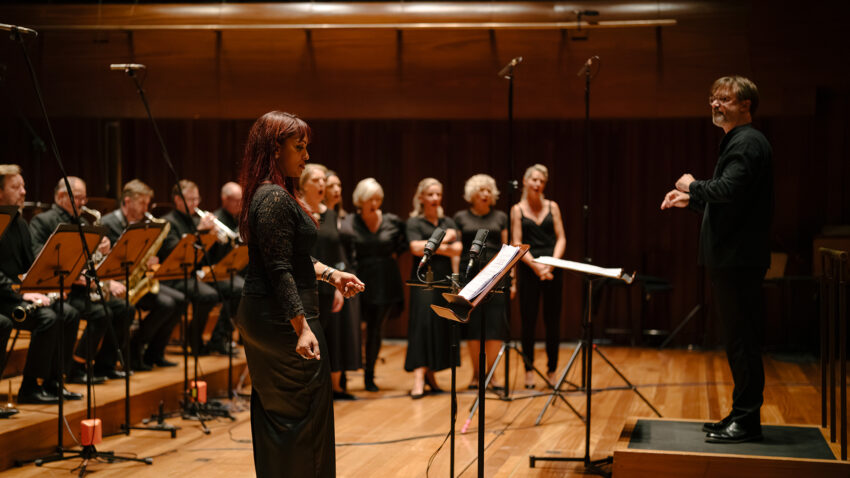 Danielle de Niese stands at a microphone in front of members of the Chorus and Orchestra of ENO. André de Ridder conducts the performance. The musicians and singers are dressed in formal black attire.
