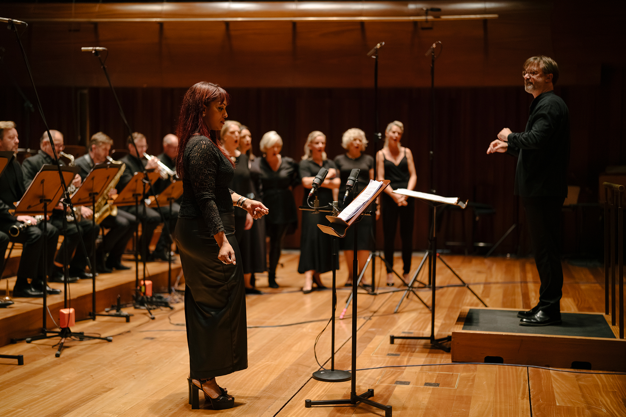 Danielle de Niese stands at a microphone in front of members of the Chorus and Orchestra of ENO. André de Ridder conducts the performance. The musicians and singers are dressed in formal black attire.
