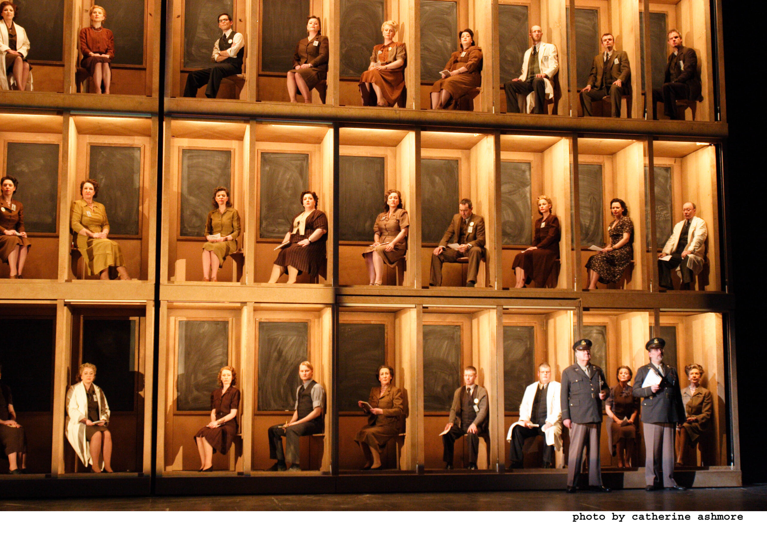 Actors in period costumes sit individually in box-like compartments stacked on stage, whilst two people in military uniforms talk at the front.