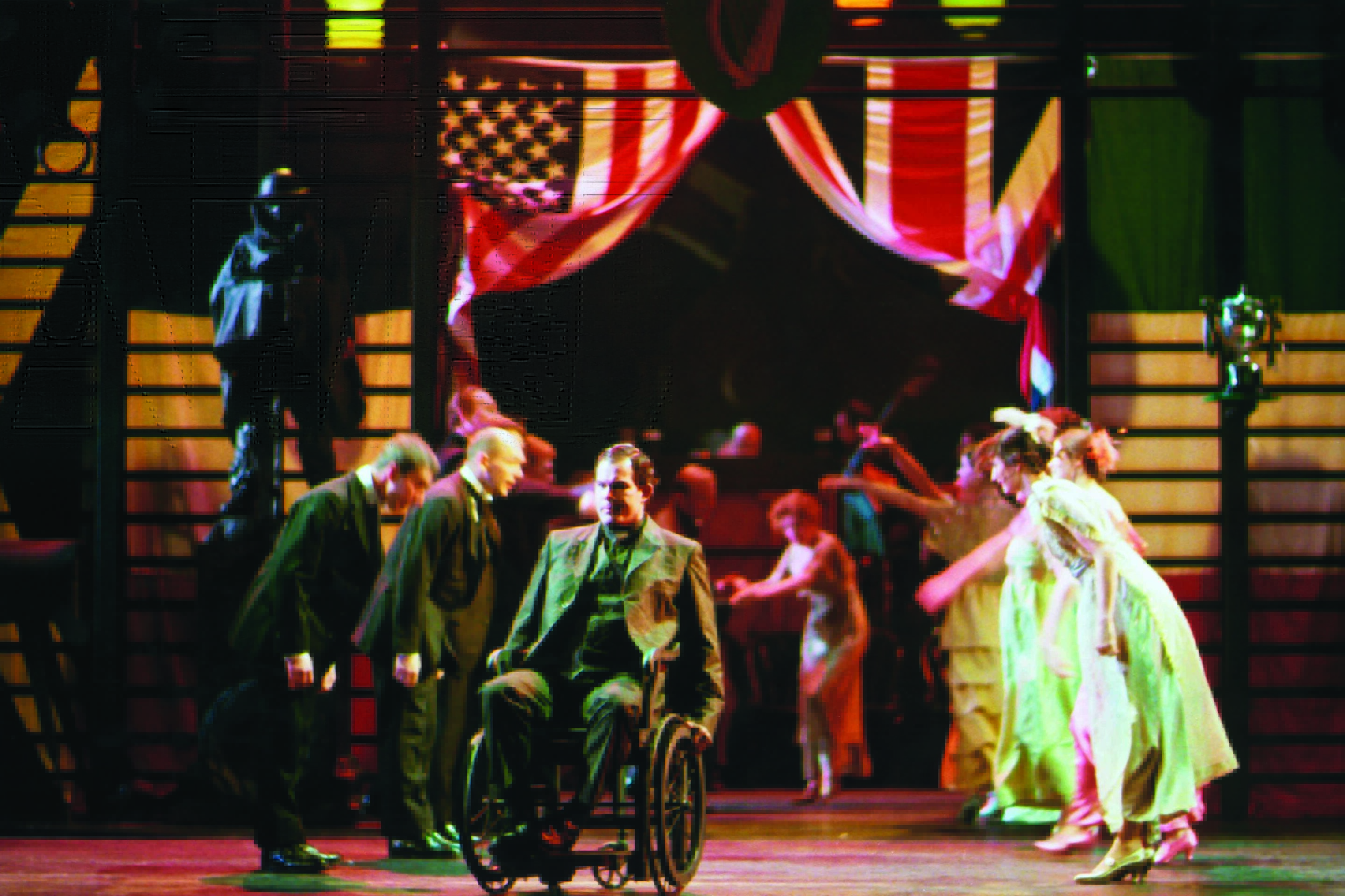 A theatrical stage scene features a man in a wheelchair at the centre, surrounded by actors in formal and vintage costumes, with an American flag backdrop and dramatic lighting.