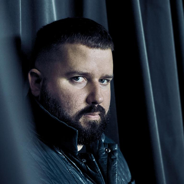 A man with a trimmed beard and short hair, wearing a dark quilted jacket, stands in front of draped dark curtains, gazing intently at the camera.