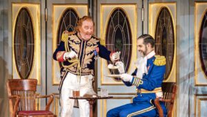 Two men in elaborate, regal costumes perform on stage. One, standing, gestures grandly while holding a spoon over a pudding; the other, seated, smiles and holds a tray. Ornate doors with oval windows form the backdrop.