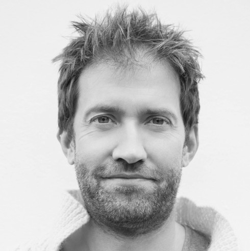Meet the Team: A man with tousled hair and a short beard, wearing a textured jumper, faces the camera and smiles slightly. The photo is in black and white with a plain background.