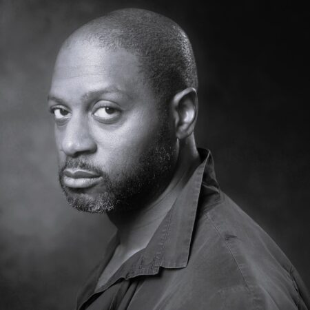 A black and white portrait of Rodney Earl Clarke, a bearded man in a collared shirt, looking seriously at the camera against a dark, softly textured background.