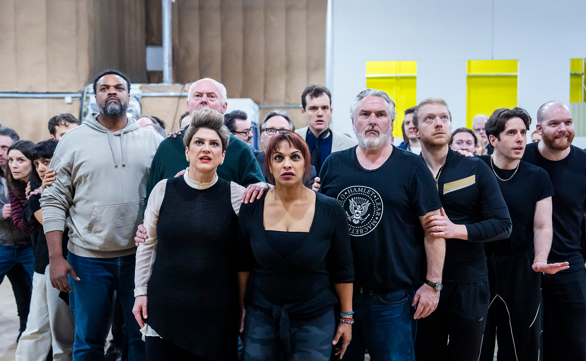 A diverse group of people stand closely together indoors, looking forward with serious, focused expressions, as if watching or anticipating something important. The background shows a warehouse or rehearsal space setting.
