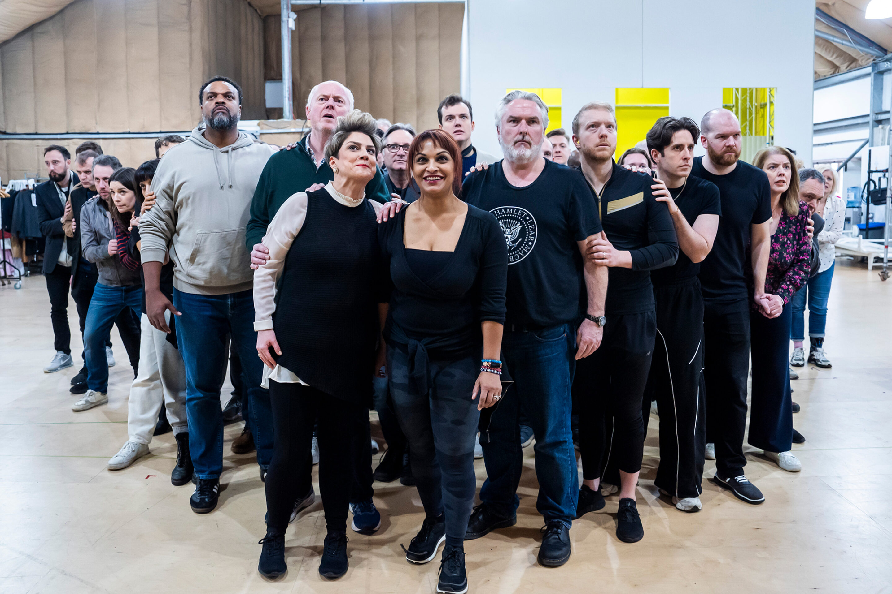 A group of people of various ages stand close together indoors, looking intently ahead. Some have their arms linked and expressions of anticipation or focus. The setting appears to be a rehearsal space or studio.