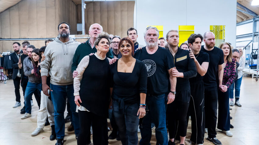 A group of people of various ages stand close together indoors, looking intently ahead. Some have their arms linked and expressions of anticipation or focus. The setting appears to be a rehearsal space or studio.