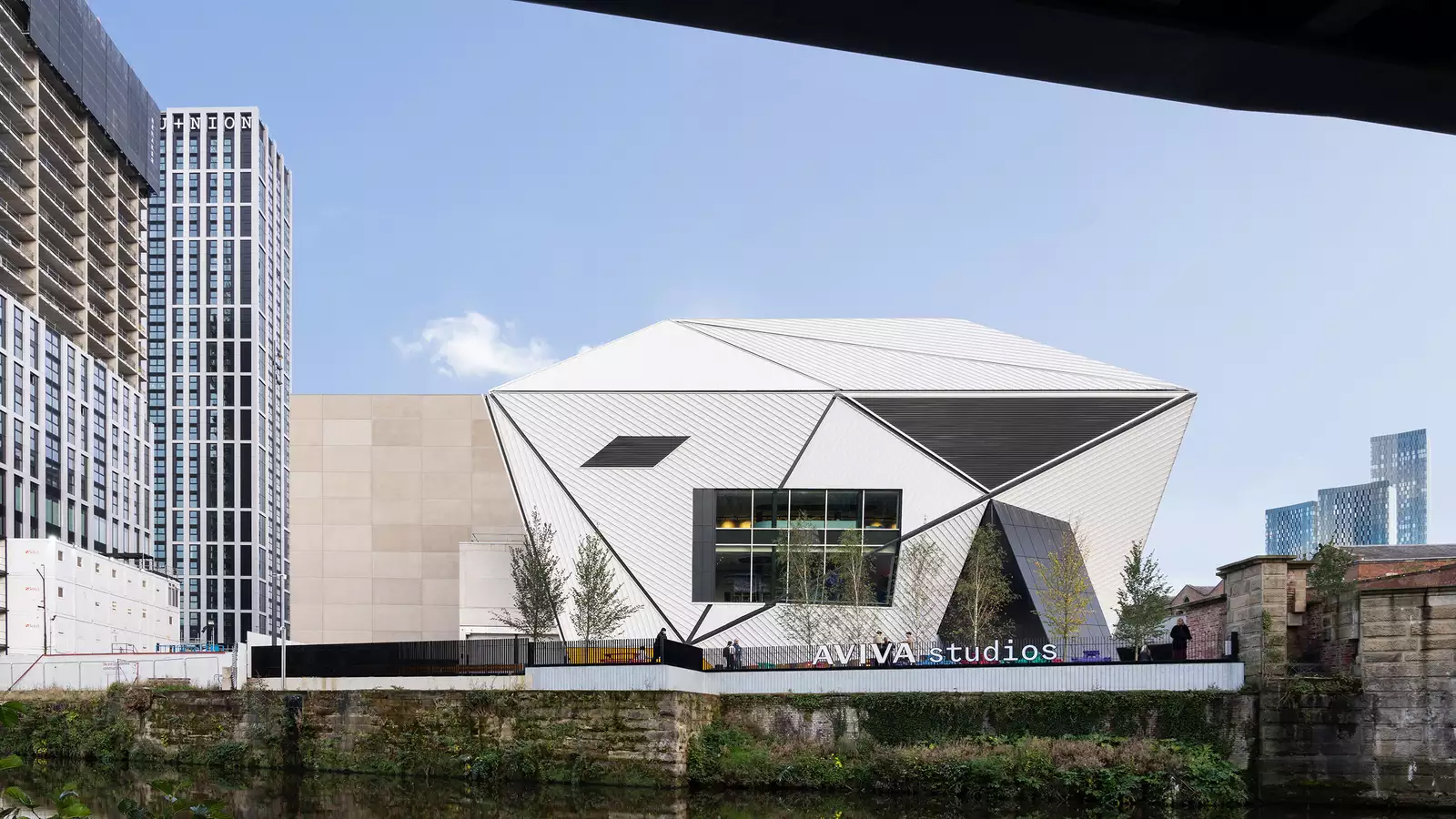 A modern, angular building with geometric white panels labelled Aviva Studios, surrounded by trees and high-rise buildings, set against a clear blue sky with a canal in the foreground.