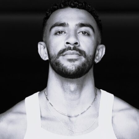 Black and white portrait of a man with short curly hair, trimmed beard, and moustache, wearing a sleeveless white vest and layered necklaces, looking confidently at the camera against a dark background.