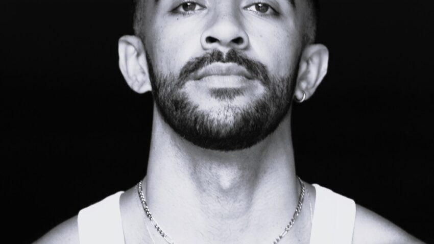 Black and white portrait of a man with short curly hair, trimmed beard, and moustache, wearing a sleeveless white vest and layered necklaces, looking confidently at the camera against a dark background.