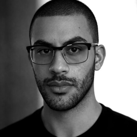 A young man with short hair, a beard, and glasses looks directly at the camera. He wears a dark shirt and the background is softly blurred in black and white.