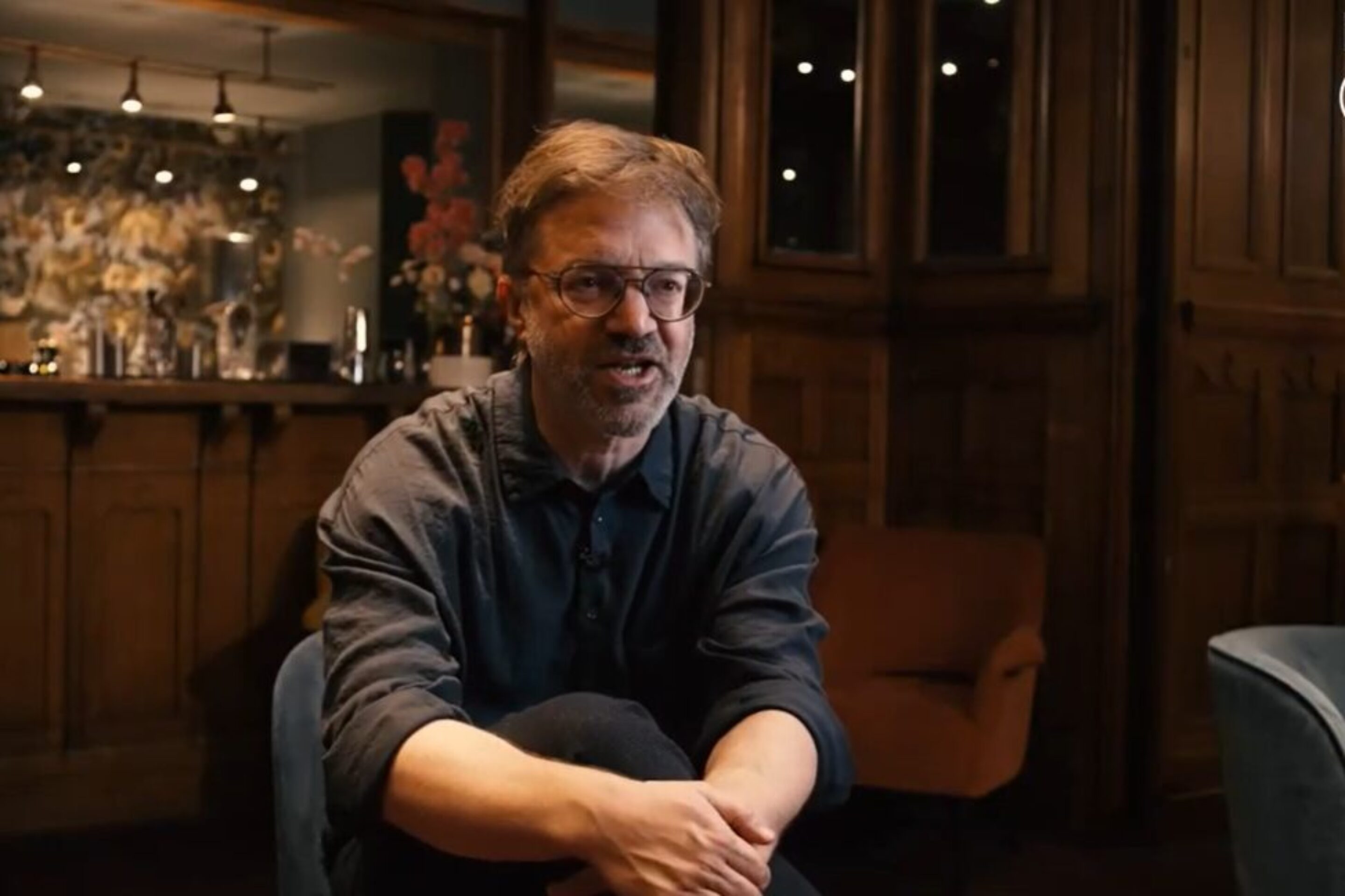 A man with glasses and a beard, resembling conductor André de Ridder, sits talking in a warmly lit room with wooden panelling and a bar in the background. He leans forward with his hands clasped. The letters ENO are in the top right corner.