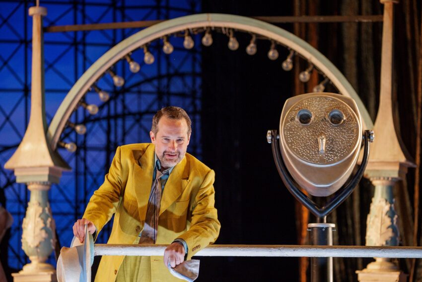 A man in a yellow suit leans on a white railing, holding a hat, with a large coin-operated binocular viewer beside him. An illuminated arch and geometric window frame the theatrical stage setting.