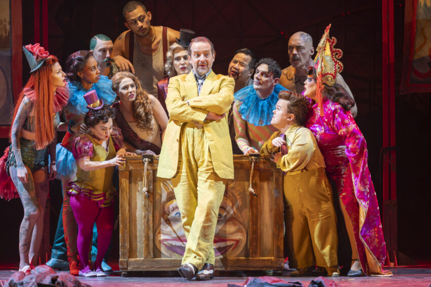 A man in a yellow suit stands confidently in front of a wooden crate, surrounded by a diverse group of circus performers in colourful costumes who are looking at him with curiosity and anticipation on a stage.
