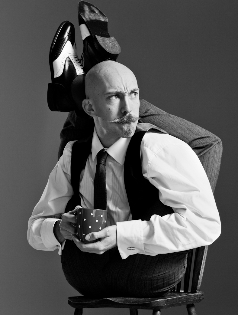 A bendy bald man in a suit sits on a chair with his legs folded over his shoulders in a contorted pose, holding a spotty mug and looking off to the side, with a serious expression and a curled moustache.