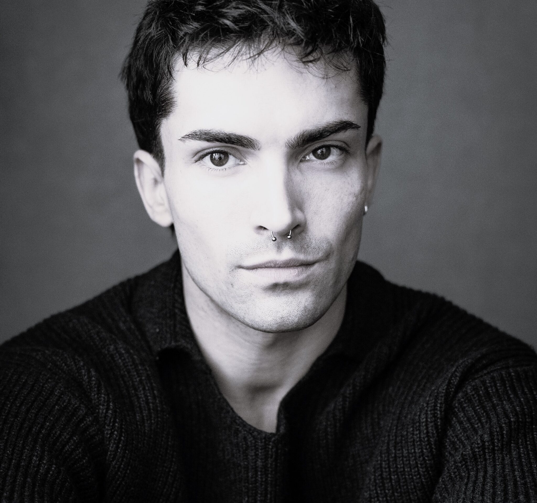 A young person with short dark hair and a septum piercing, wearing a dark jumper, looks directly at the camera with a neutral expression against a plain background.