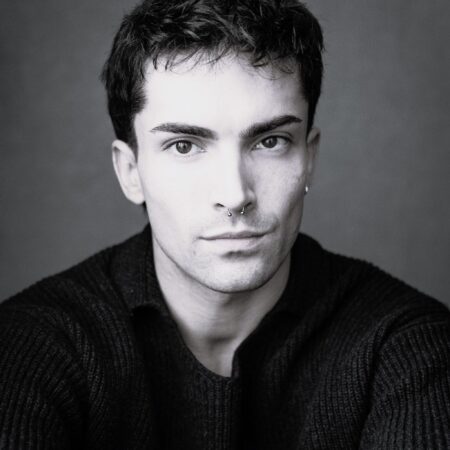 A young person with short dark hair and a septum piercing, wearing a dark jumper, looks directly at the camera with a neutral expression against a plain background.