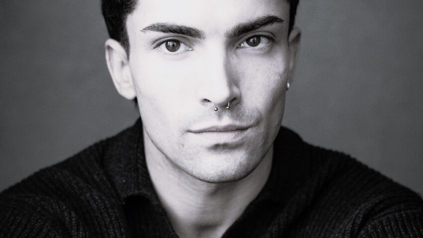 A young person with short dark hair and a septum piercing, wearing a dark jumper, looks directly at the camera with a neutral expression against a plain background.
