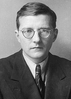 Black and white portrait of Russian composer Dmitri Shostakovich, a man with short hair and round glasses, wearing a suit and tie, looking directly at the camera with a neutral expression.