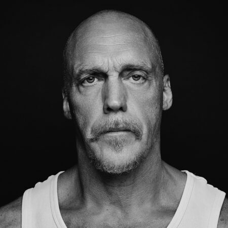 A bald man with a moustache and goatee, resembling Duncan Meadows, stares seriously at the camera. He is wearing a white vest; the dark background and black-and-white photo emphasise his facial features.