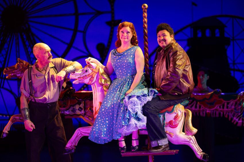 Three actors on stage in a theatrical scene: a woman in a blue polka dot dress sits on a carousel horse, flanked by two men—one in a brown leather jacket sitting behind her, the other standing beside the horse.