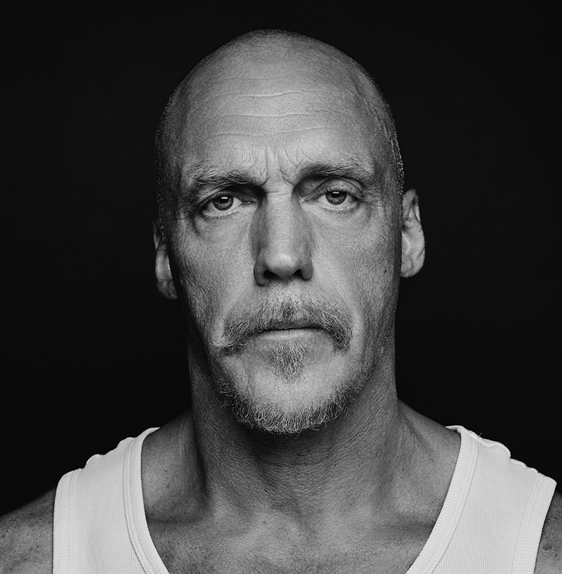 A bald man with a moustache and goatee, resembling Duncan Meadows, stares seriously at the camera. He is wearing a white vest; the dark background and black-and-white photo emphasise his facial features.
