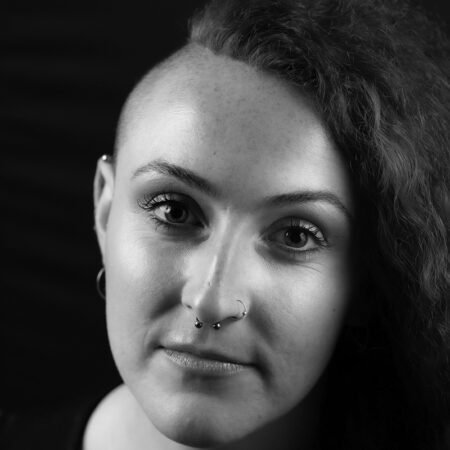 Jessica Catherine, with one side of her head shaved and the other side sporting curly hair, looks at the camera. She has a septum and nose ring, hoop earrings, and a neutral expression in this black-and-white photo.