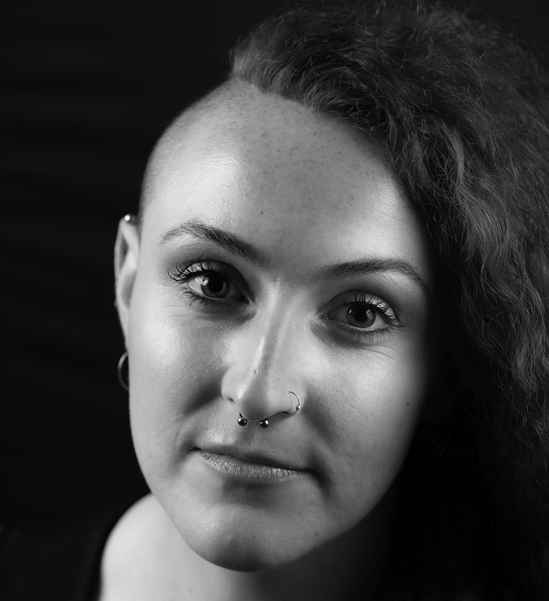 Jessica Catherine, with one side of her head shaved and the other side sporting curly hair, looks at the camera. She has a septum and nose ring, hoop earrings, and a neutral expression in this black-and-white photo.