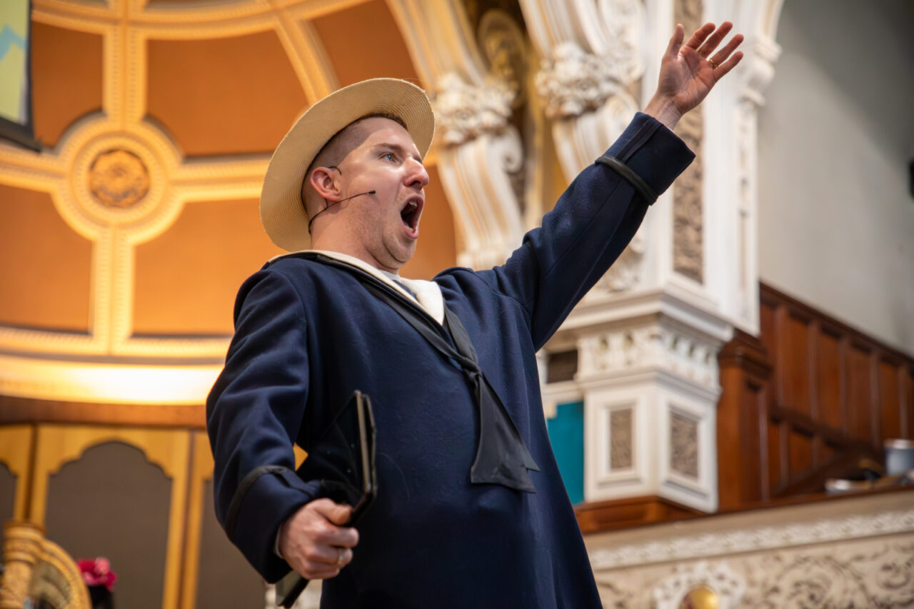 A performer in a sailor costume sings passionately on stage, raising one arm and holding a prop, as ornate architectural details shine in the background. For our school newsletter interview, Michael Jones talks about working on Finish This!.