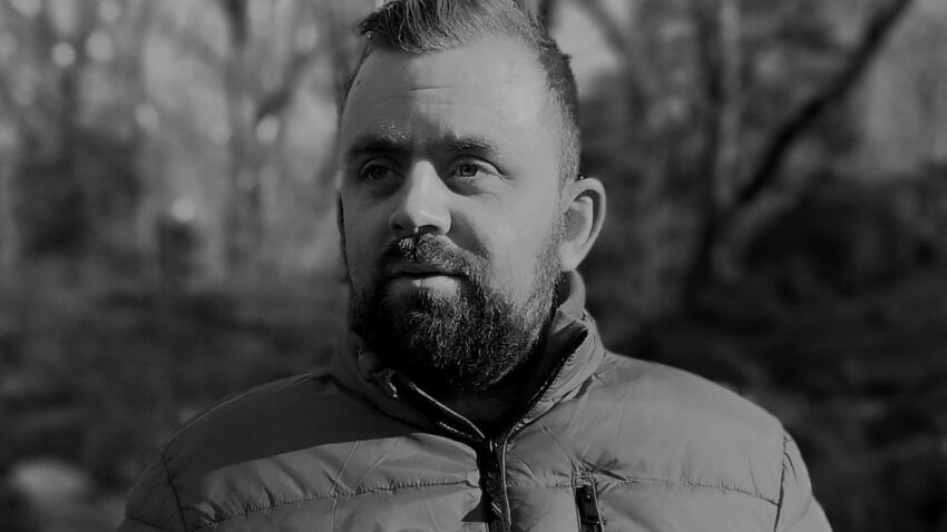 A bearded man wearing a padded jacket stands outdoors, looking slightly to the side. The background is blurred with trees, and the image is in black and white.