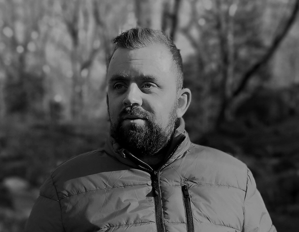 A bearded man wearing a padded jacket stands outdoors, looking slightly to the side. The background is blurred with trees, and the image is in black and white.