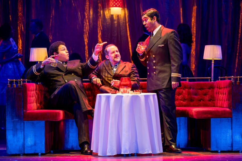 Three men in formal attire sit and stand around a small round table with drinks, set on a stage with red velvet sofas, illuminated lamps, and a shiny gold curtain backdrop.