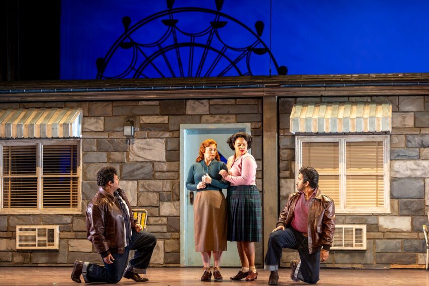 Four actors perform on stage in front of a stone house set. Two women stand by the door, one comforting the other. Two men in brown jackets kneel on either side, facing them. A blue backdrop with a Ferris wheel silhouette is behind.