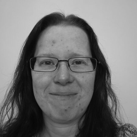 Laura Sprake, with long dark hair and glasses, smiles gently at the camera. The black and white photo features a plain, light-coloured background.
