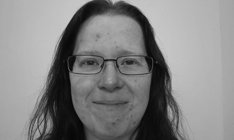 Laura Sprake, with long dark hair and glasses, smiles gently at the camera. The black and white photo features a plain, light-coloured background.