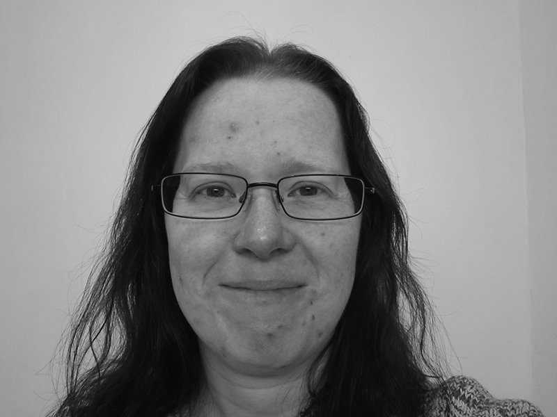 Laura Sprake, with long dark hair and glasses, smiles gently at the camera. The black and white photo features a plain, light-coloured background.