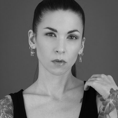 Leah Debrincat, with long dark hair tied back, hoop earrings, and a sleeveless top, looks seriously at the camera. She shows off her tattoos as she touches her shoulder with one hand in this striking black and white image.
